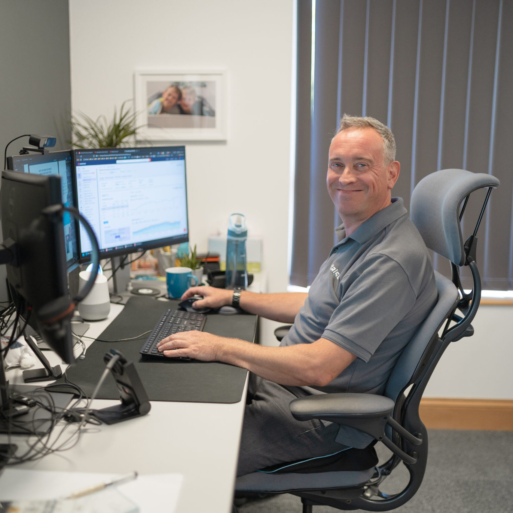 Man sitting at office desk working on two monitors, smiling at the camera, with gray blinds behind.