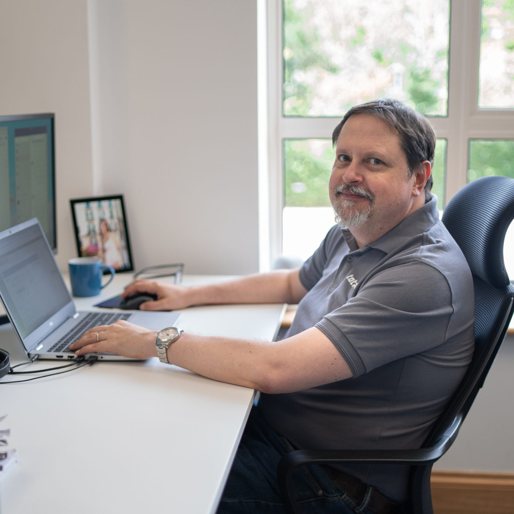 Man sitting at a desk working on a laptop in a bright office with a window and framed photo.