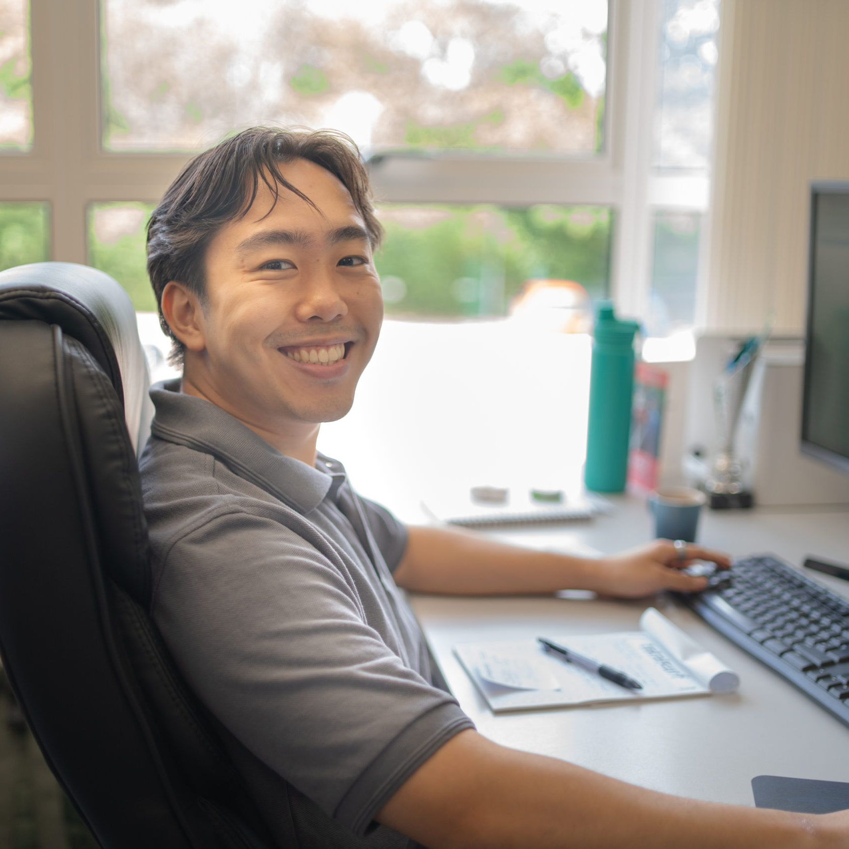 Man sitting at a desk, smiling at the camera, with a computer, notepad, and pen in front of him.