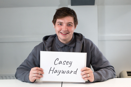 Person sitting at a table holding a sign that reads "Casey Hayward" and smiling at the camera.