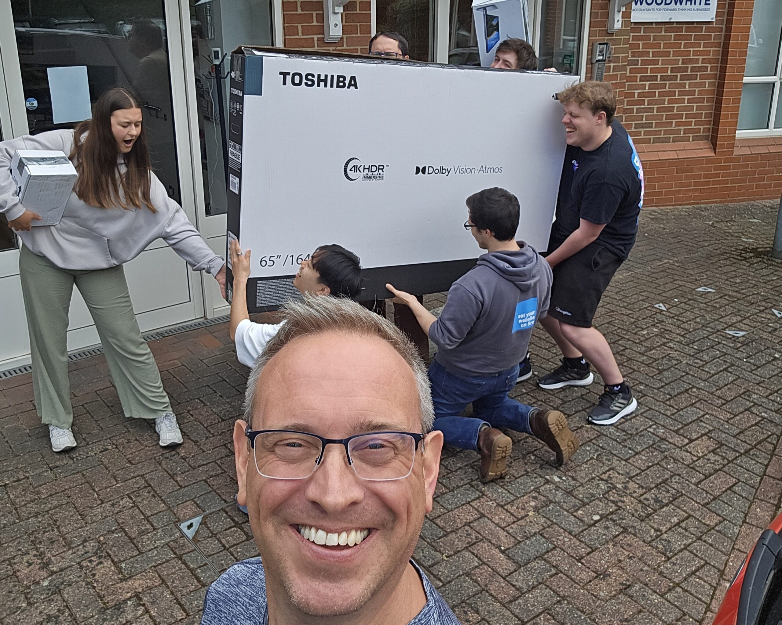 Group of people carrying a large Toshiba TV box outside, while one person takes a selfie in front.