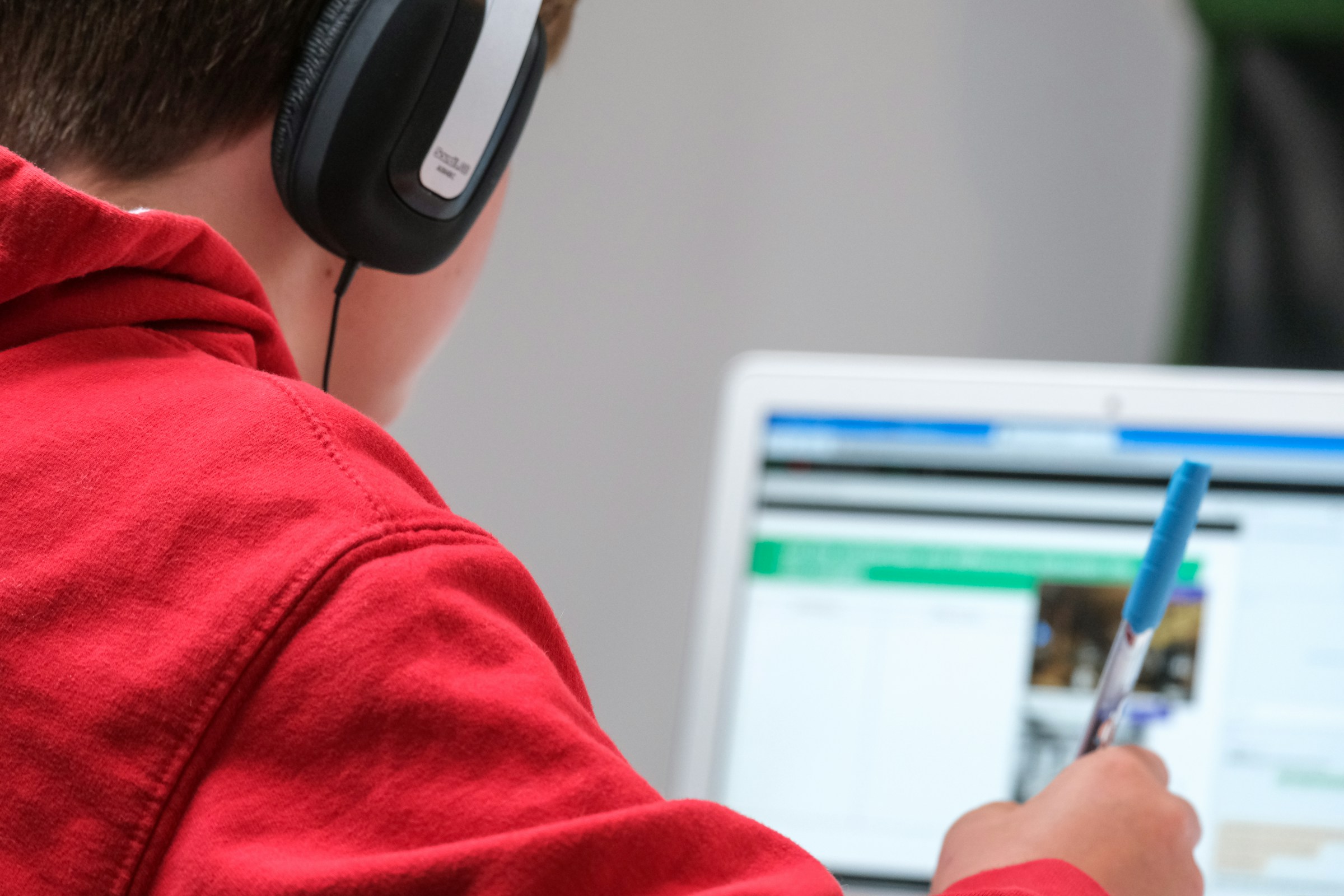 Person in red hoodie wearing headphones studies at a computer, holding a pen in one hand.