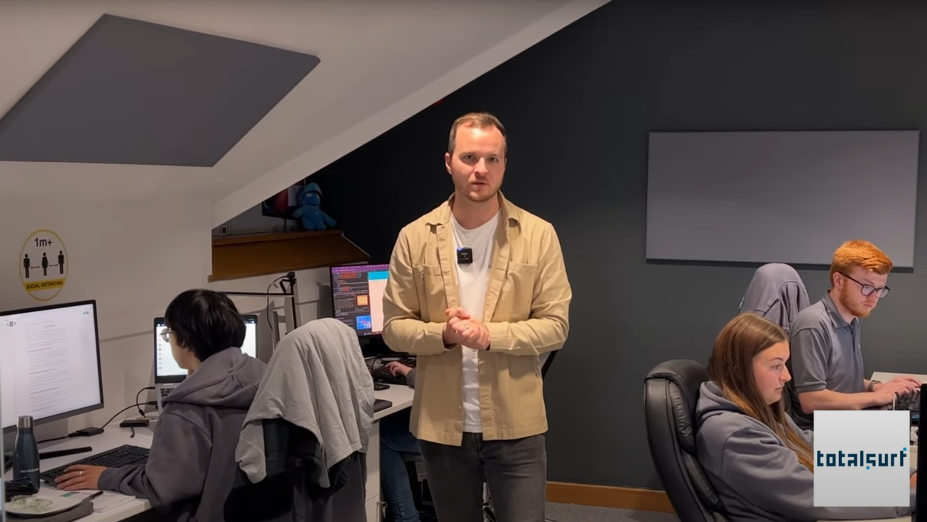 Five people work at computers in an office; one person stands in the center facing the camera.