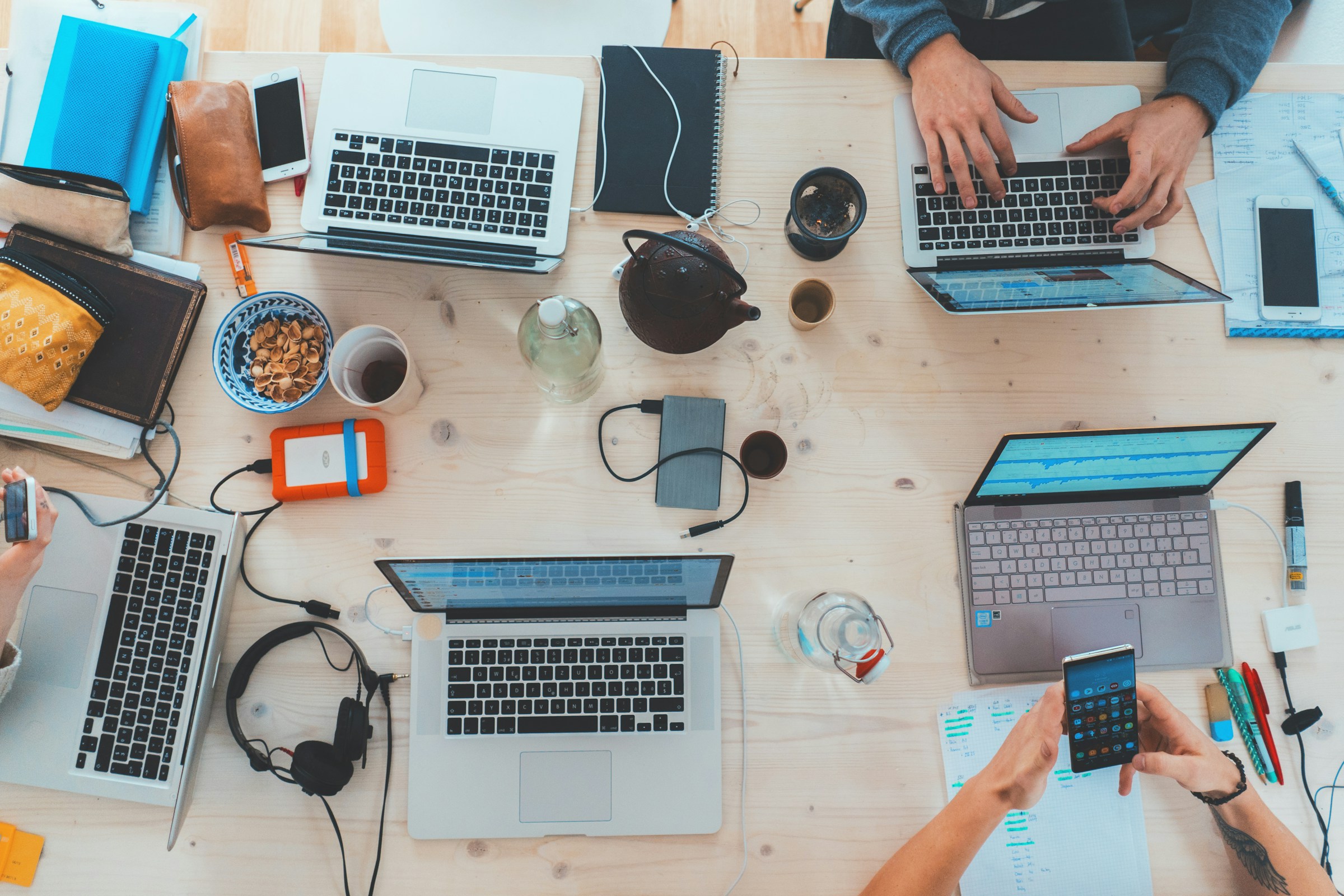 Four people working at a table with laptops, notebooks, snacks, and electronic devices.
