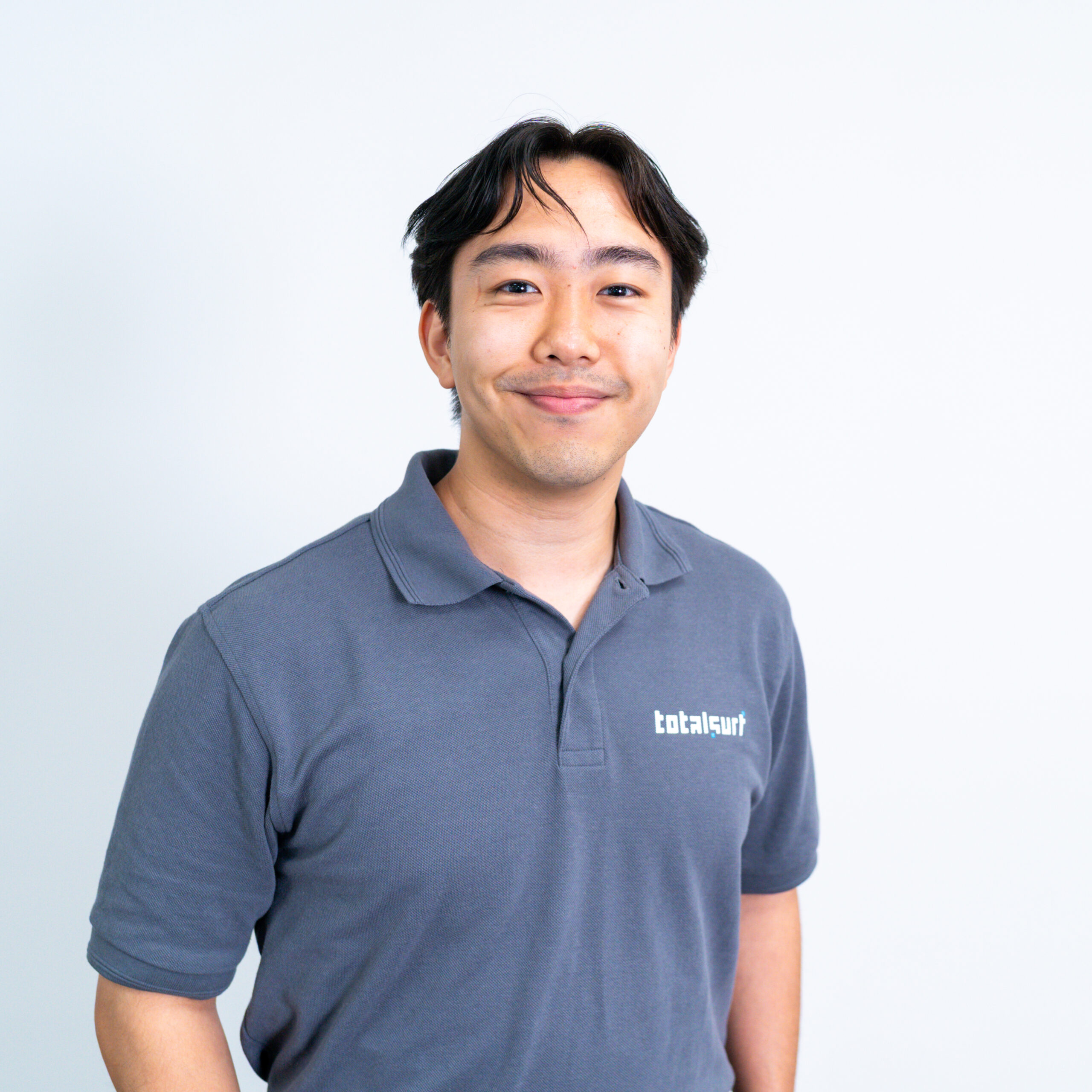 Person in a grey polo shirt with "totalsurf" logo stands against a plain white background, smiling.