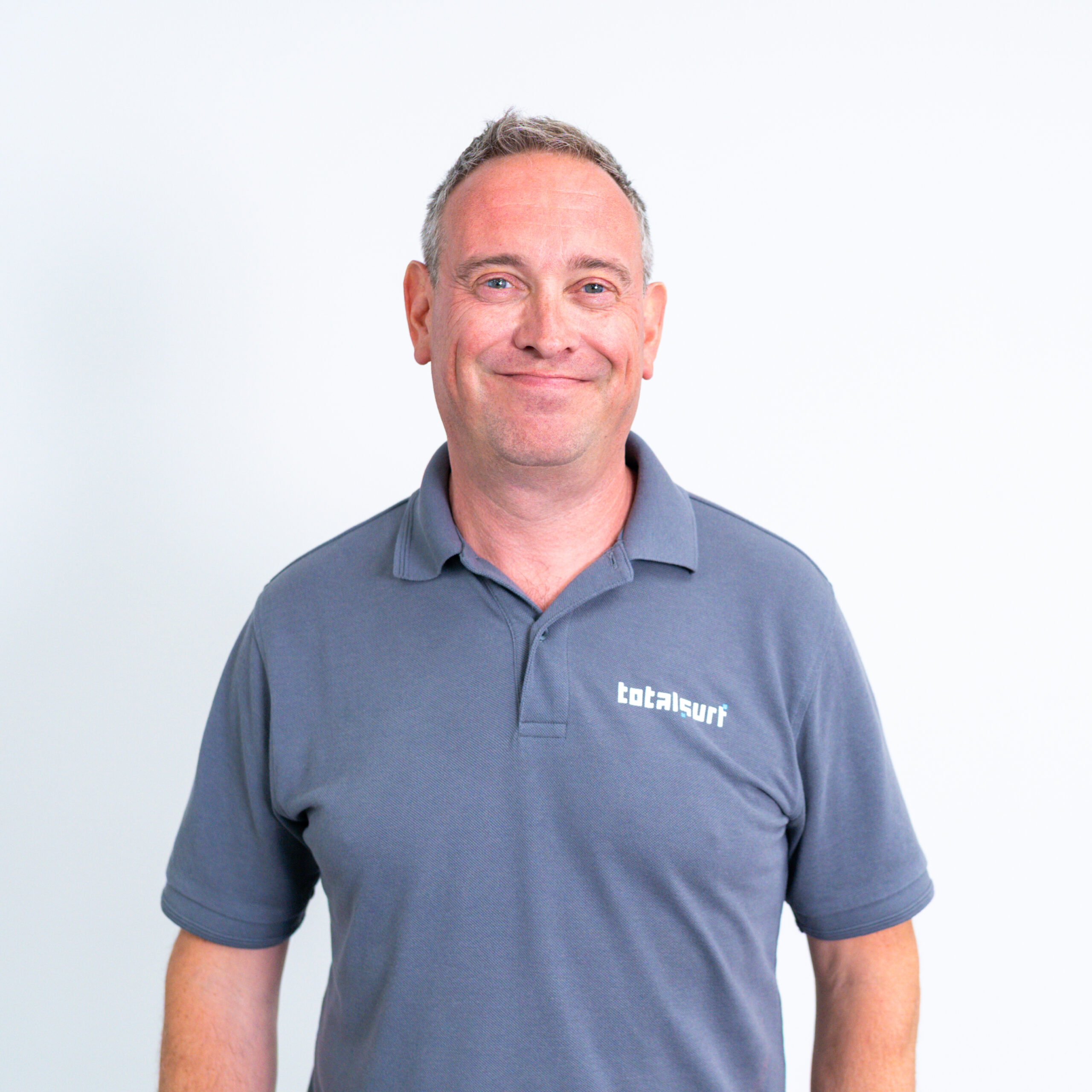 A man in a grey "totalsurf" polo shirt stands against a plain white background, smiling.