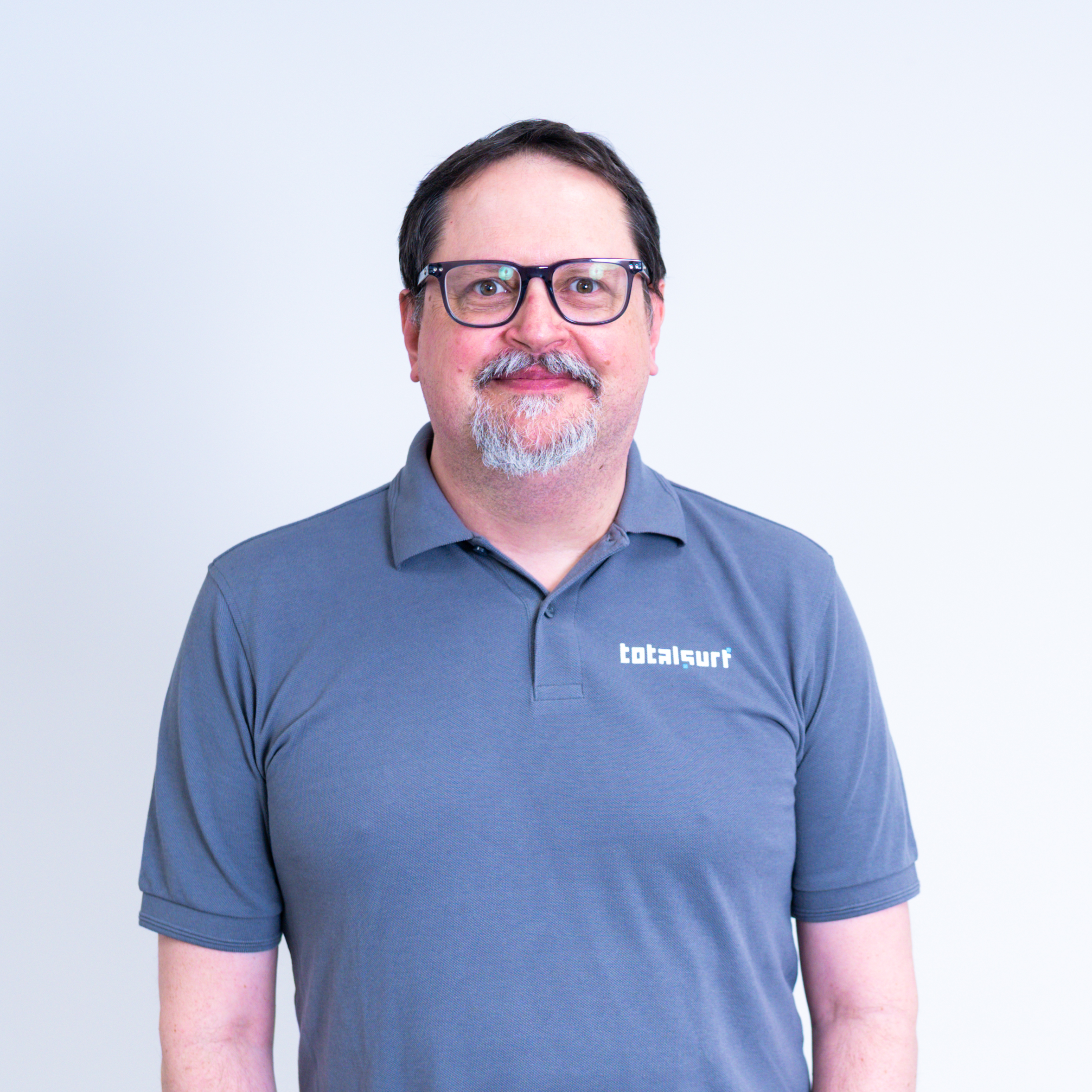 Man with glasses wearing a gray "totalsurf" polo shirt stands against a plain light background.