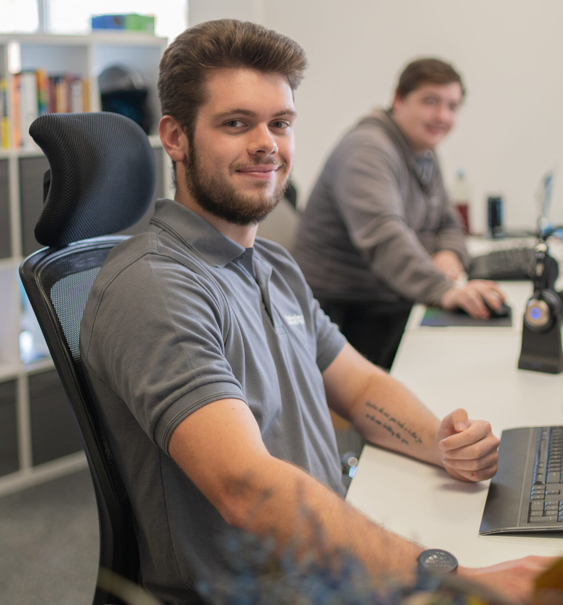 Man in a gray polo shirt sits at a desk in an office, smiling toward the camera.