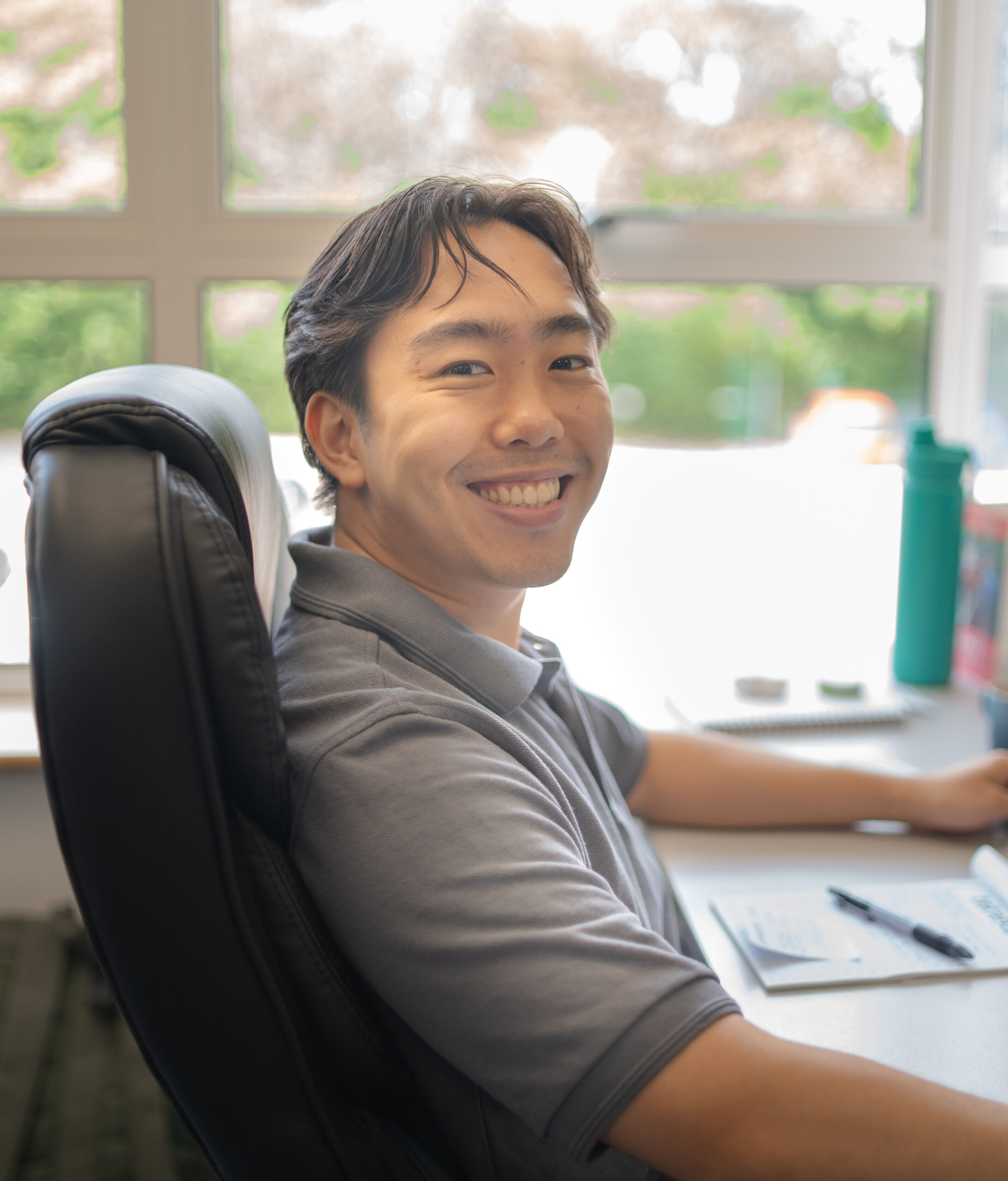 A person sitting at a desk, smiling at the camera, with papers and a water bottle nearby.