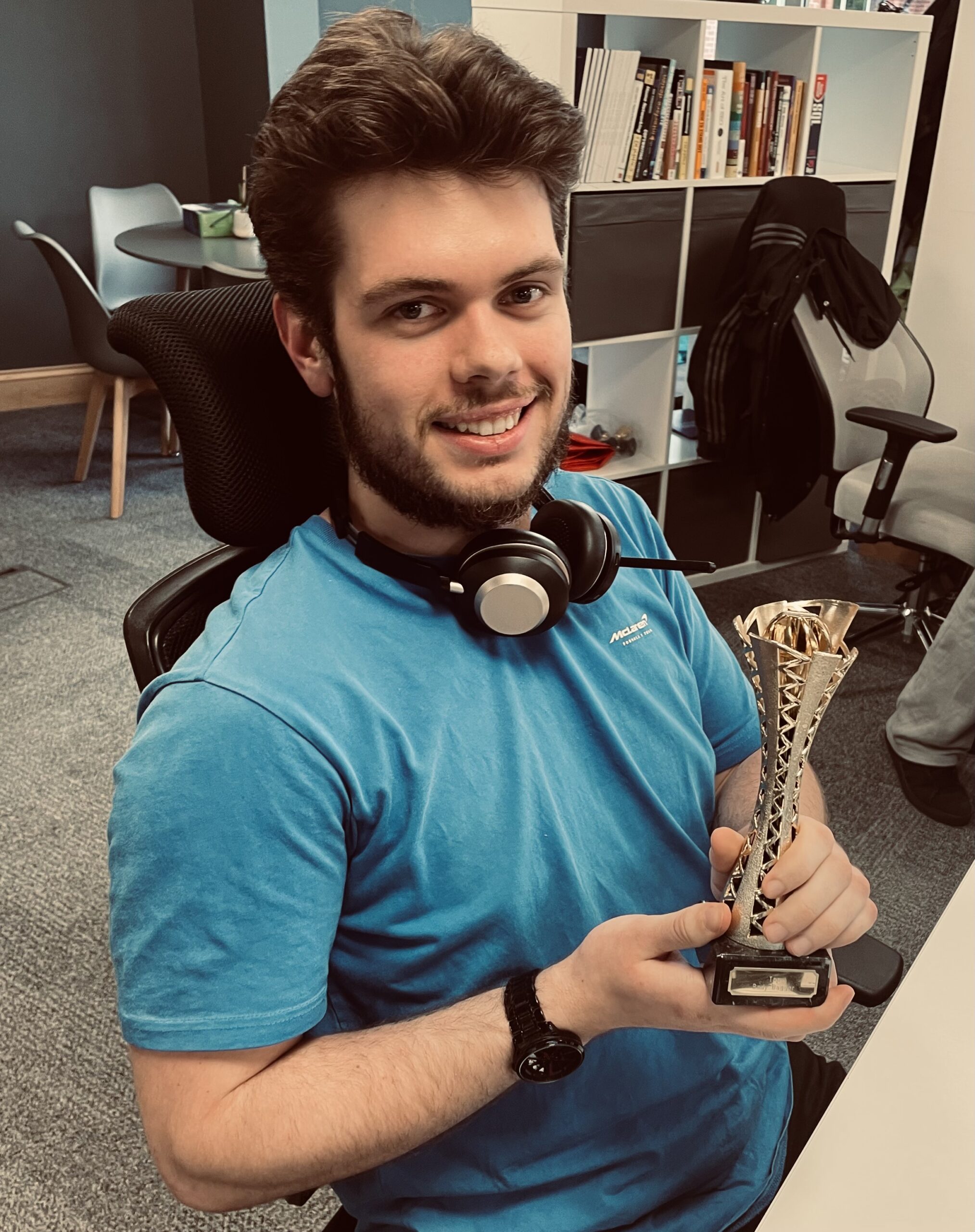A man in a blue shirt sits at a desk, smiling and holding a small trophy in his hands.