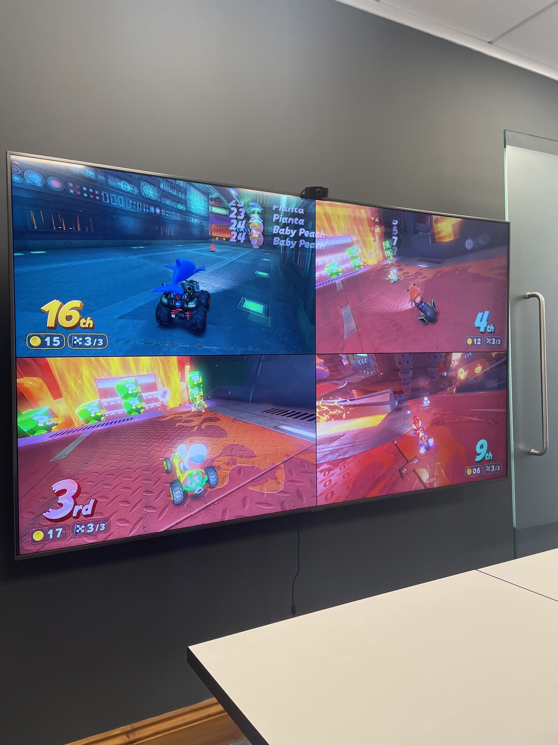 A four-player Mario Kart race is shown on a TV screen with a green hue, mounted on a gray wall.