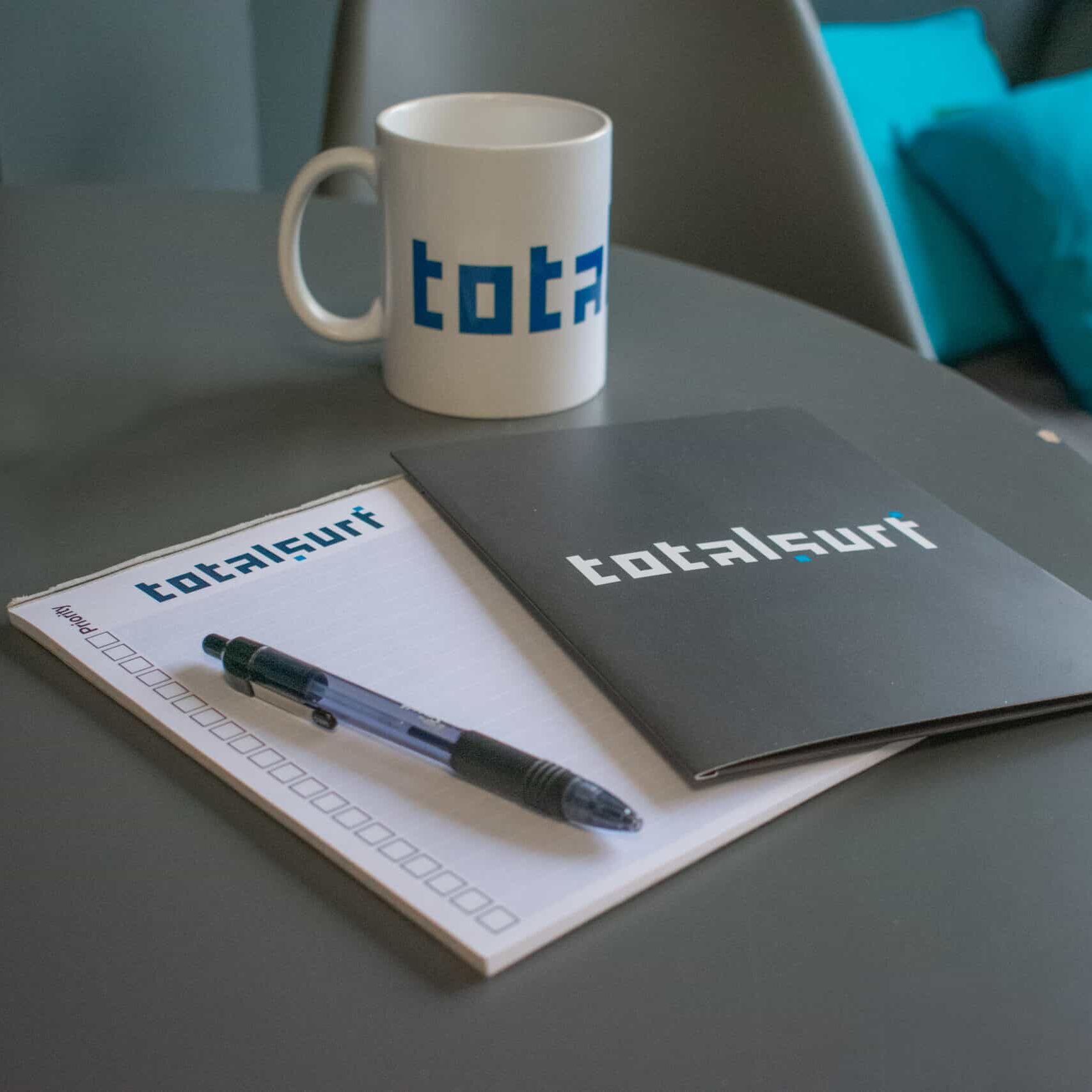 A mug, notebook, pen, and pad with "totalsurf" branding on a gray table.