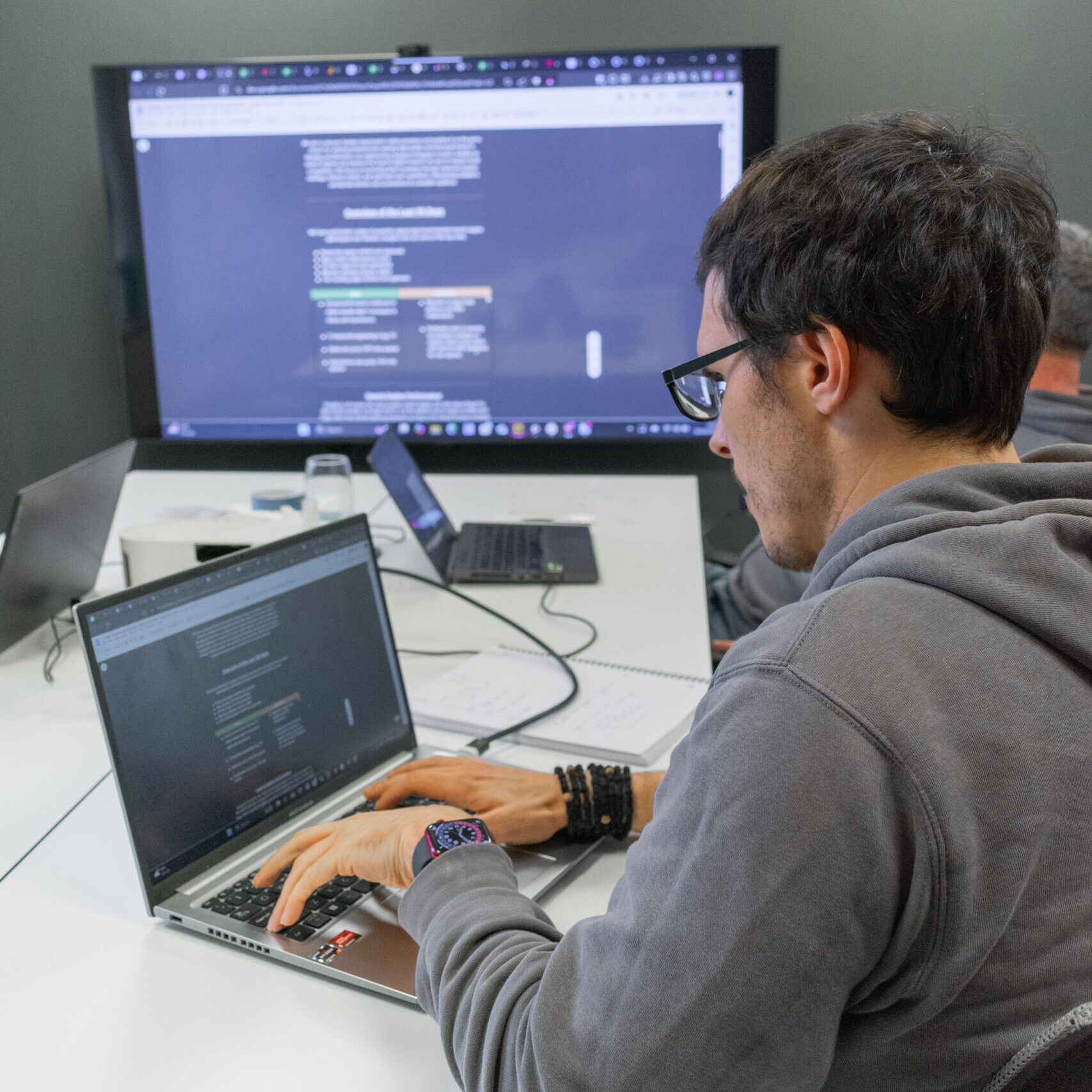 Person in glasses typing on a laptop at a desk, with code displayed on a large screen behind.